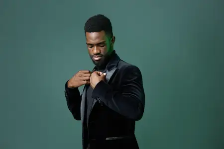 Уверенный в себе мужчина в чёрном смокинге поправляет галстук на фоне ярко-зелёного фона, излучая утончённость и стиль