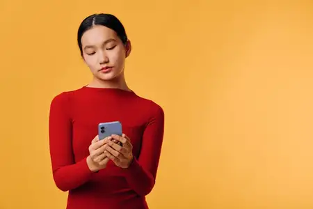 Молодая женщина в красном топе держит смартфон, глядя на экран, изолированная на оранжевом фоне, сосредоточенная на телефоне для общения, цифрового использования, набора текста, просмотра…