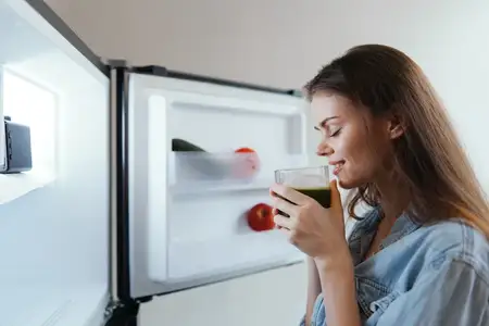 Молодая женщина с длинными каштановыми волосами улыбается, смакуя зелёный смузи перед холодильником, в котором стоят свежие фрукты и овощи