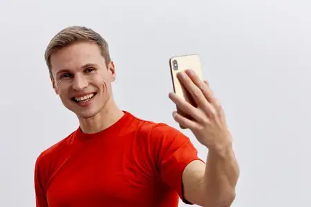 Улыбающийся мужчина делает селфи на телефон в умной повседневной красной футболке, изолированный на однотонном нейтральном фоне
