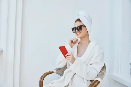 Женщина с телефоном в белом халате сидит на балконе в отеле