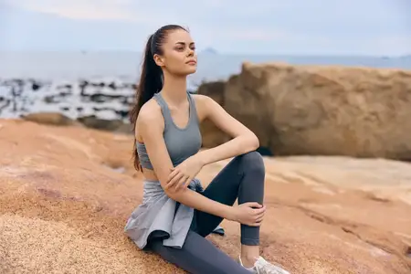 Подтянутая молодая женщина в спортивной одежде мирно сидит на каменистом пляже, принимая осознанность и благополучие