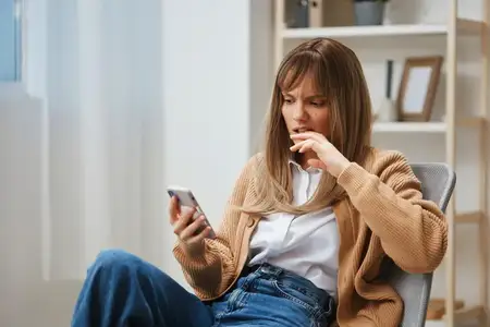 Сбитая с толку и несчастная молодая светловолосая девушка в тёплом свитере читает плохие новости, используя телефон, сидя в кресле в современном домашнем интерьере