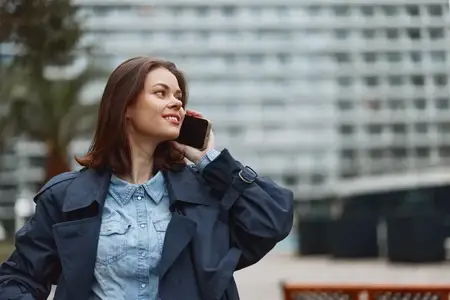 Веселая молодая женщина в синем пальто пользуется смартфоном на улице в городской среде с размытым современным зданием на заднем плане