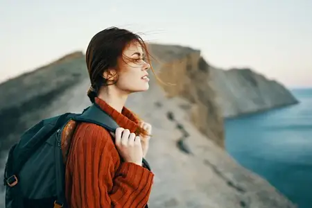 Женщина-туристка с рюкзаком в свитере отдыхает в горах рядом с морем и смотрит в небо