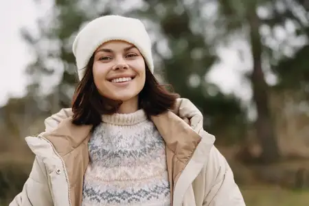 Уличный портрет улыбающейся женщины в тёплом пальто и вязаной шапке-бини, наслаждающейся прохладным днём на природе, излучающей комфорт, моду и позитивную энергию