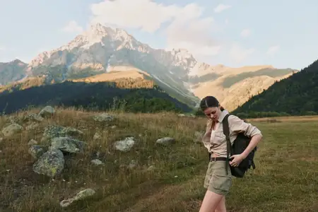 Молодая женщина в бежевой рубашке и хаки-шортах несет небольшой рюкзак, пересекая травянистую тропу в горной долине под ясным небом, излучая спокойствие и любопытство