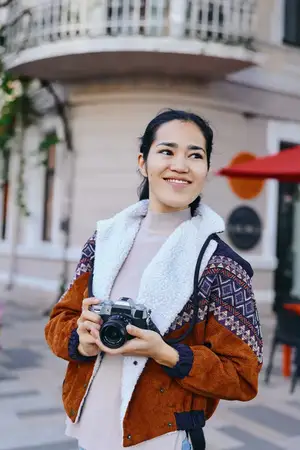Уличная сцена: радостная женщина с фотоаппаратом в тёплой куртке снимает моменты на залитой солнцем городской улице