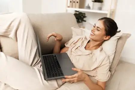 Счастливая женщина работает из дома за ноутбуком, улыбается и наслаждается комфортом гостиной