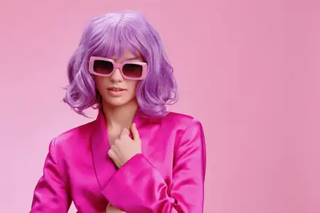 Женщина с фиолетовыми волосами в солнцезащитных очках и розовом костюме уверенно стоит на розовом фоне