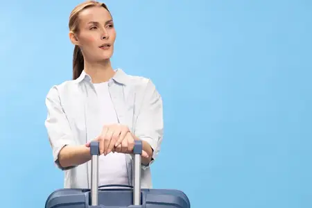 Молодая женщина, держащаяся за ручку чемодана, смотрит в сторону на синем фоне