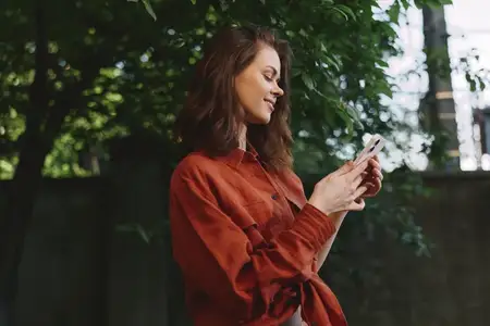 Она стоит рядом с бетонной стеной на фоне пышной зелени, одетая в длиннорукавную блузу ржавого цвета, наслаждаясь тенью и спокойным моментом повседневной жизни в городском парке