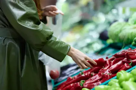 Женщина в продуктовом магазине выбирает свежие овощи для приготовления ужина дома, держа в руках красный перец во время покупок на рынке