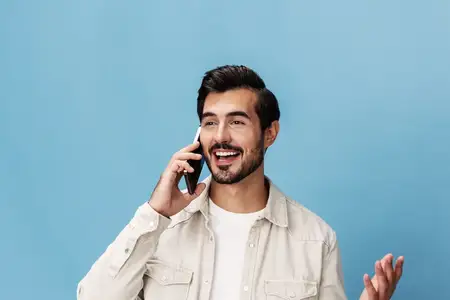 Крупный план портрета брюнета, который разговаривает по телефону с другом, улыбается, обнажая зубы, на синем фоне, одет в белую футболку и джинсы