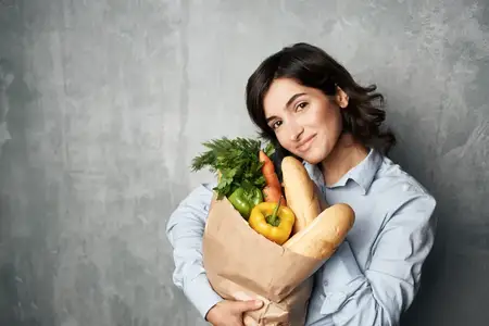 Брюнетка с пакетом продуктов в синей рубашке на фоне овощей и тёмного фона