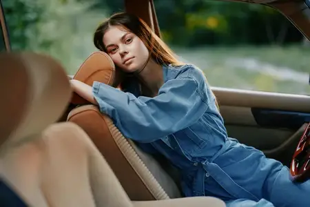 Молодая женщина в джинсовом наряде, прислонившись к автомобилю, излучает спокойствие и безмятежность на фоне размытого уличного пейзажа, отражая момент умиротворения