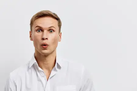Удивленный молодой светлокожий мужчина с открытым ртом и широко раскрытыми глазами, в белой рубашке, смотрит в камеру, изолирован на однотонном светлом фоне, выражая шок или изумление