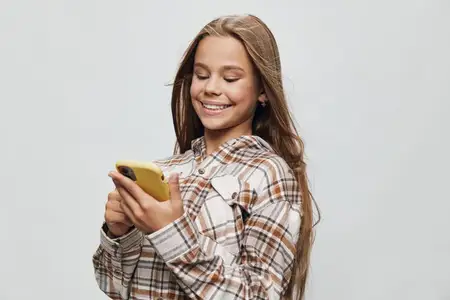 Счастливая девушка-подросток с длинными волосами пользуется смартфоном, одетая в клетчатую рубашку, стоит на белом фоне