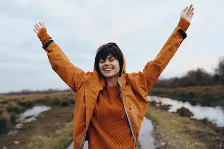 Женщина на открытом воздухе с широкой улыбкой, подняв руки вверх, стоит на тропинке