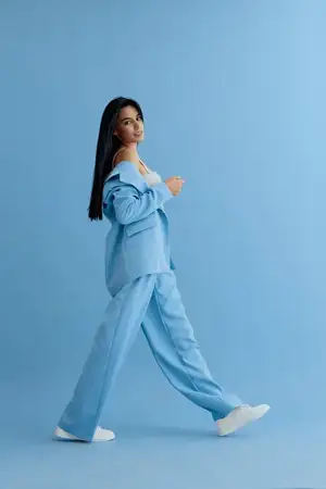 Молодая женщина уверенно идет в синем oversized костюме и белых кроссовках, ее длинные темные волосы естественно развеваются, на фоне сплошного светло-голубого фона для современного модного образа