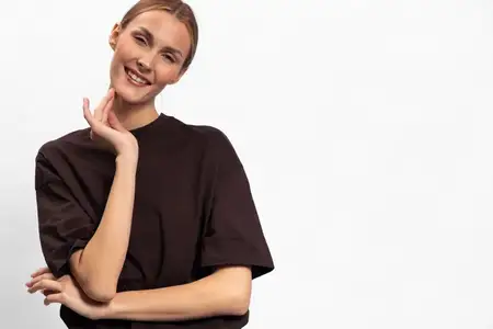 Счастливая молодая женщина в черной футболке стоит на белом фоне, улыбается и нежно касается рукой лица