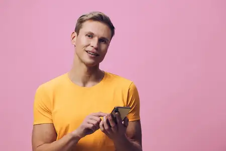 Улыбающийся мужчина пользуется мобильным телефоном, одетый в повседневную оранжевую футболку, изолированный на розовом фоне, студийный портрет