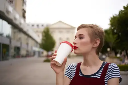 Женщина с короткими волосами на улице, в очках, идёт со стаканом напитка в руке