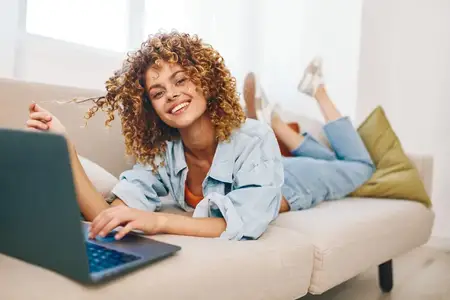 Улыбающаяся женщина печатает на ноутбуке, наслаждаясь работой фрилансера в уютном домашнем офисе