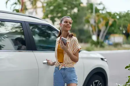Весёлая молодая женщина стоит рядом с белым автомобилем, держа свой смартфон