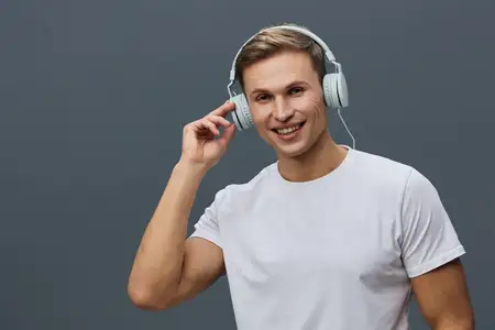 Счастливый молодой человек в накладных наушниках и белой футболке улыбается и слушает музыку на фоне однотонного серого фона