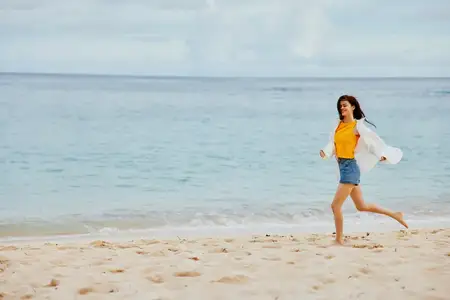Спортсменка бежит по пляжу в летней одежде по песку: на ней жёлтая футболка и джинсовые шорты, белая рубашка развевается, волосы летят на ветру, на заднем плане виден океан
