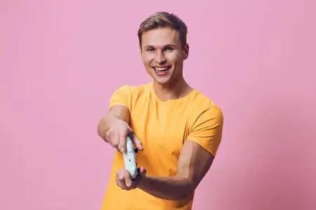 Улыбающийся молодой человек в желтой футболке держит геймпад, играет в видеоигры, демонстрирует волнение и веселье, изолирован на розовом фоне