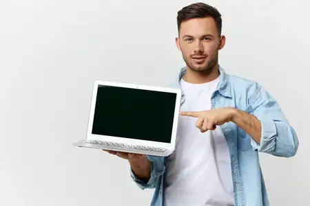 Серьезный, дружелюбный, загорелый, красивый ИТ-специалист в повседневной простой футболке указывает пальцем на ноутбук, позирует изолированно на белом студийном фоне