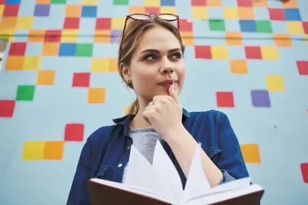 Студентка в рубашке и очках держит в руках книгу на улице возле граффити на стене
