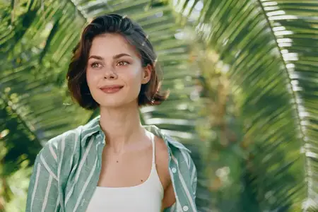 Уличное изображение женщины, улыбающейся в тропическом саду, повседневная одежда, яркий солнечный свет, расслабленная поза, естественный фон, создающий привлекательное настроение и жизнерадостное…