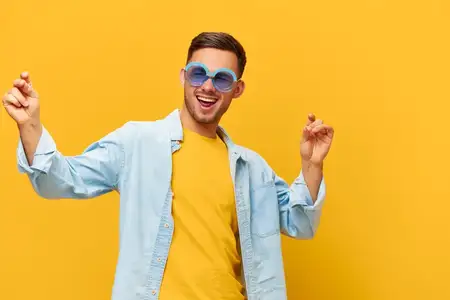 Танцующий радостный загорелый красивый мужчина в синей рубашке и модных солнцезащитных очках поднимает руки вверх, позируя изолированно на оранжево-желтом студийном фоне