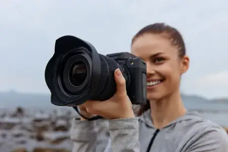 Веселая молодая женщина с камерой фотографирует живописный пляж, одетая в серую толстовку