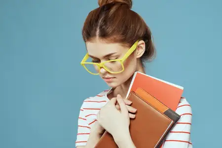 Студентка с книгами в руках на синем фоне, в жёлтых очках и с модельной причёской, кадрированный вид
