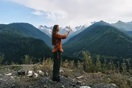 Сцена на открытом воздухе с женщиной, фотографирующей обширный горный хребет, оранжевый топ, пересеченная местность, вечнозеленые деревья и ясное небо, передающие приключение и природу