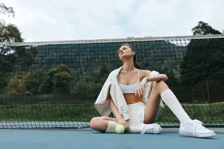 Юная спортивная женщина уверенно позирует на теннисном корте в спортивном наряде, излучая счастье и расслабление в естественном зеленом окружении