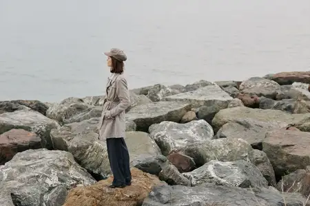 Женщина в пальто и шляпе стоит одна на камнях у моря, глядя вдаль