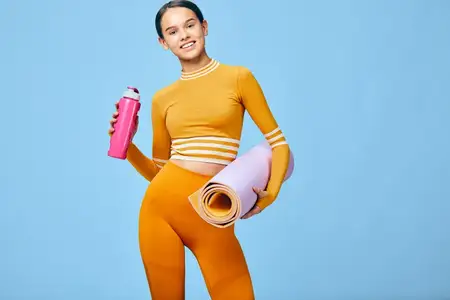 Спортивная женщина-атлет в ярком костюме с ковриком и бутылкой, готовая к упражнениям и оздоровительным тренировкам, энергичная поза для фитнеса, тренажерного зала и домашних тренировок