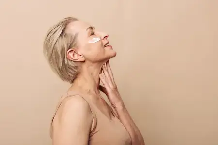 Счастливая женщина средних лет касается рукой шеи, одетая в бежевый топ и с патчами для глаз