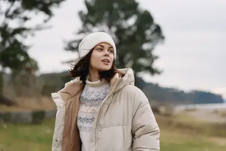 Уличный портрет женщины в стеганой куртке и уютном вязаном свитере, шапке-бини, смотрящей в небо на фоне спокойного пейзажа и приглушенных цветов вокруг