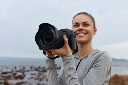 Улыбающаяся молодая женщина в серой толстовке держит камеру у моря, олицетворяя уверенность и страсть к фотографии на фоне природы