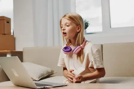 Светловолосый ребенок в розовых наушниках выразительно поет, пользуясь ноутбуком в помещении