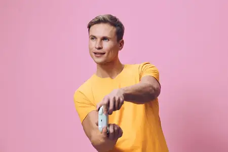 Счастливый мужчина держит смартфон, улыбается и делает селфи в повседневной жёлтой футболке на розовом студийном фоне