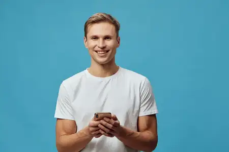 Уверенный молодой человек с яркой улыбкой держит мобильный телефон, стоя на однотонном синем фоне