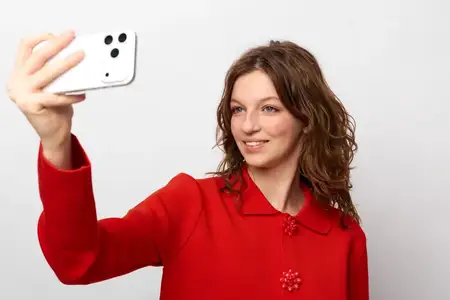 Улыбающаяся девушка в красном свитере держит телефон для селфи на простом белом фоне
