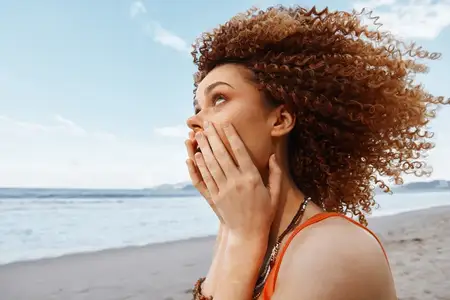 Счастье на пляже: молодая привлекательная женщина с вьющимися афро-волосами смеётся и наслаждается летним отдыхом на свежем воздухе